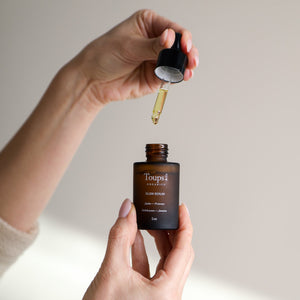 A person holds a dropper above an open bottle labeled Toups and Co Organics Glow Serum, displaying this healing serum against a neutral background.