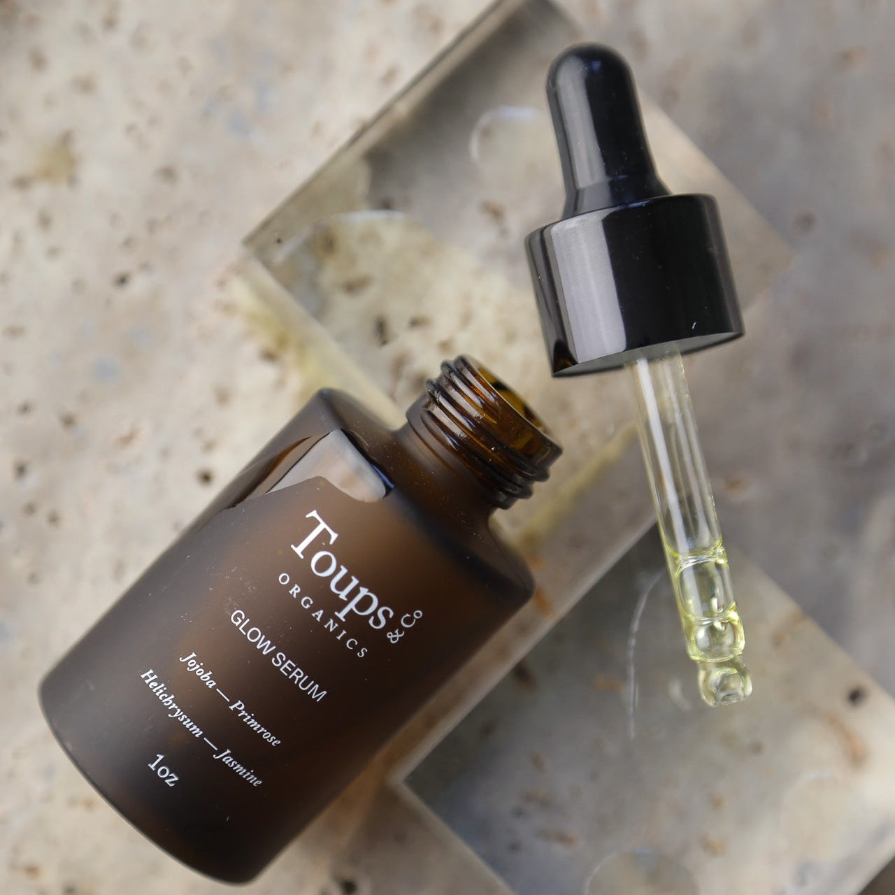 A brown glass bottle labeled Toups and Co Organics Glow Serum sits on a stone surface, featuring an open dropper filled with yellow liquid. This serum is crafted with vitamin-rich oils designed to nourish the skin and enhance its natural glow.