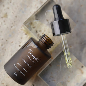 A brown glass bottle labeled Toups and Co Organics Glow Serum sits on a stone surface, featuring an open dropper filled with yellow liquid. This serum is crafted with vitamin-rich oils designed to nourish the skin and enhance its natural glow.
