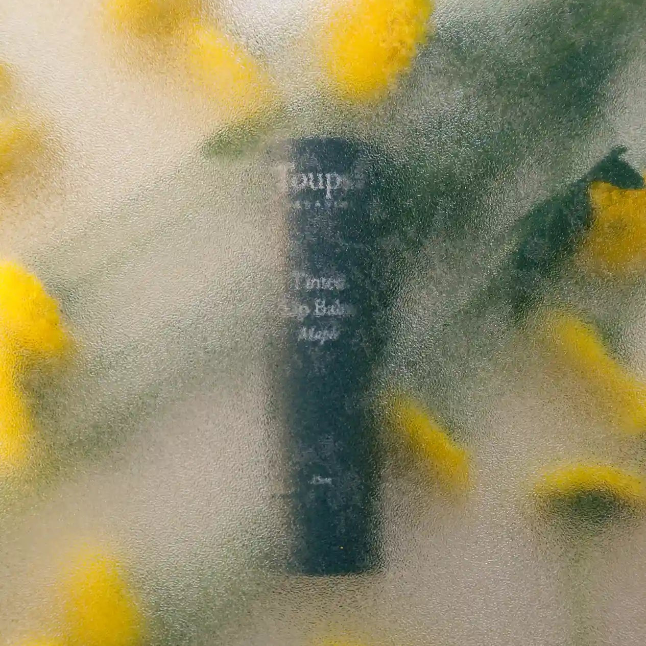 A dark cylindrical bottle of New Product by Toups and Co Organics is visible through frosted glass, with blurred yellow flowers and green leaves in the background.