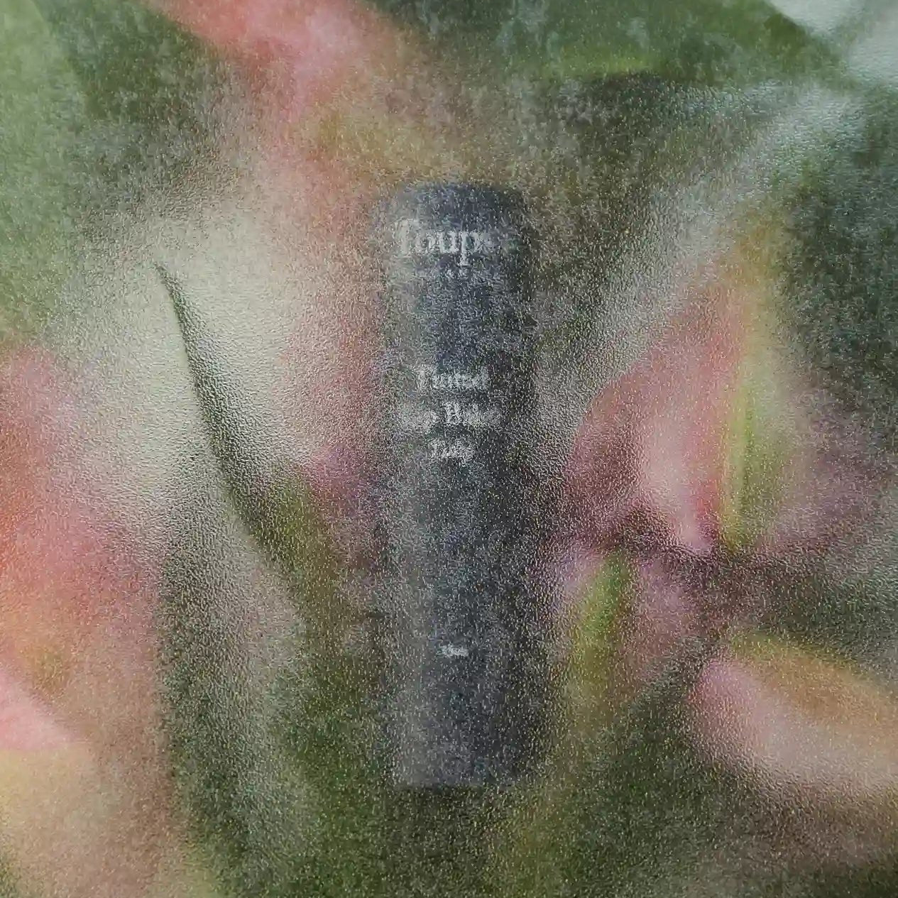 A classic black cylindrical New Product by Toups and Co Organics with white text is visible through a frosted surface, surrounded by blurred pink and green shapes that resemble non-toxic plants or flowers.
