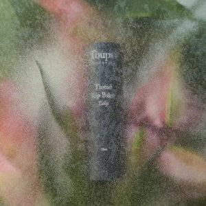 A classic black cylindrical New Product by Toups and Co Organics with white text is visible through a frosted surface, surrounded by blurred pink and green shapes that resemble non-toxic plants or flowers.