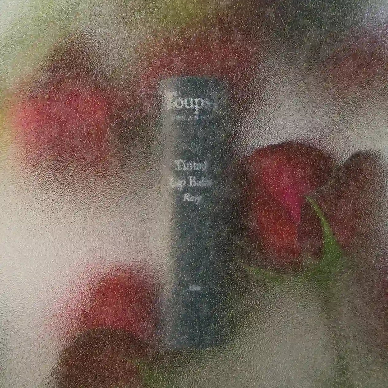 A classic dark cylindrical New Product from Toups and Co Organics is partially hidden by frosted glass, with red roses in the background, emphasizing its elegant and non-toxic appeal.