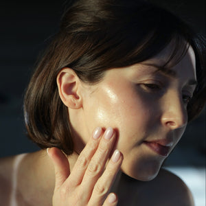 A woman with short brown hair touches her cheek, showing clear skin and a natural glow in soft lighting.