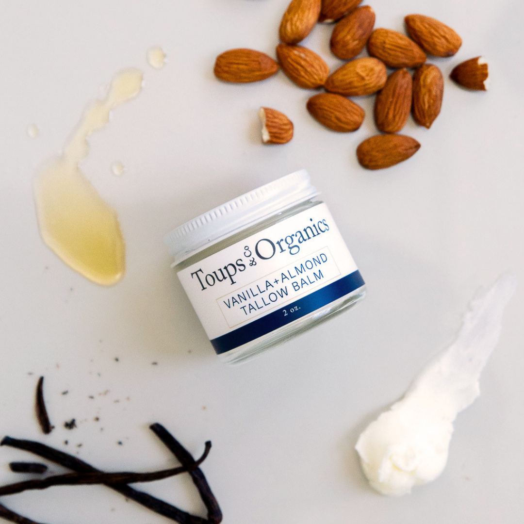 A jar of Toups and Co Organics Tallow Balm - Vanilla + Almond, crafted with nourishing grass-fed tallow for exceptional skin hydration, sits on a white surface alongside whole almonds, vanilla beans, oil, and cream.