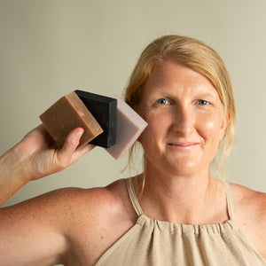A woman holds three Toups and Co Organics Vanilla Peppermint Exfoliating Body Bars—brown, black, and lavender—next to her ear and smiles at the camera.