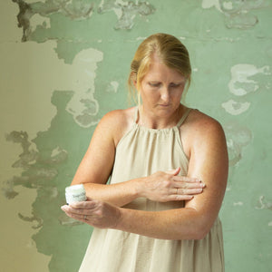 A woman applies Toups and Co Organics Non Toxic Starter Set cream to her arm while holding the small jar in her other hand, standing in front of a textured green wall.