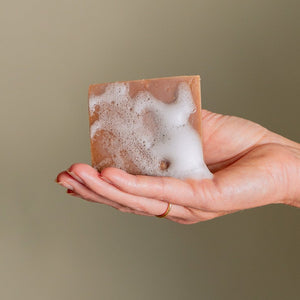 A hand adorned with a gold ring holds the Vanilla Peppermint Exfoliating Body Bar from Toups and Co Organics, covered in white foam against a neutral background.