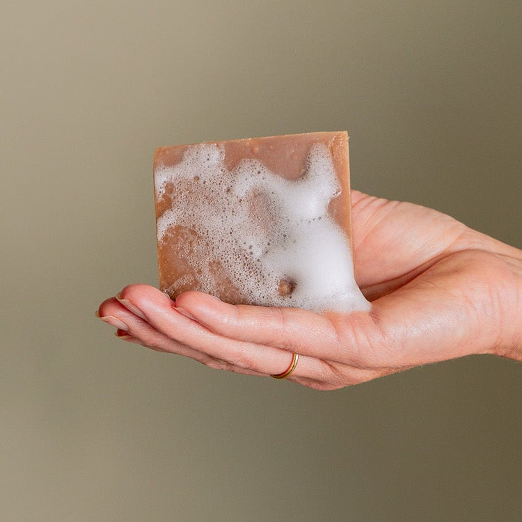 A hand adorned with a gold ring holds the Vanilla Peppermint Exfoliating Body Bar from Toups and Co Organics, covered in white foam against a neutral background.