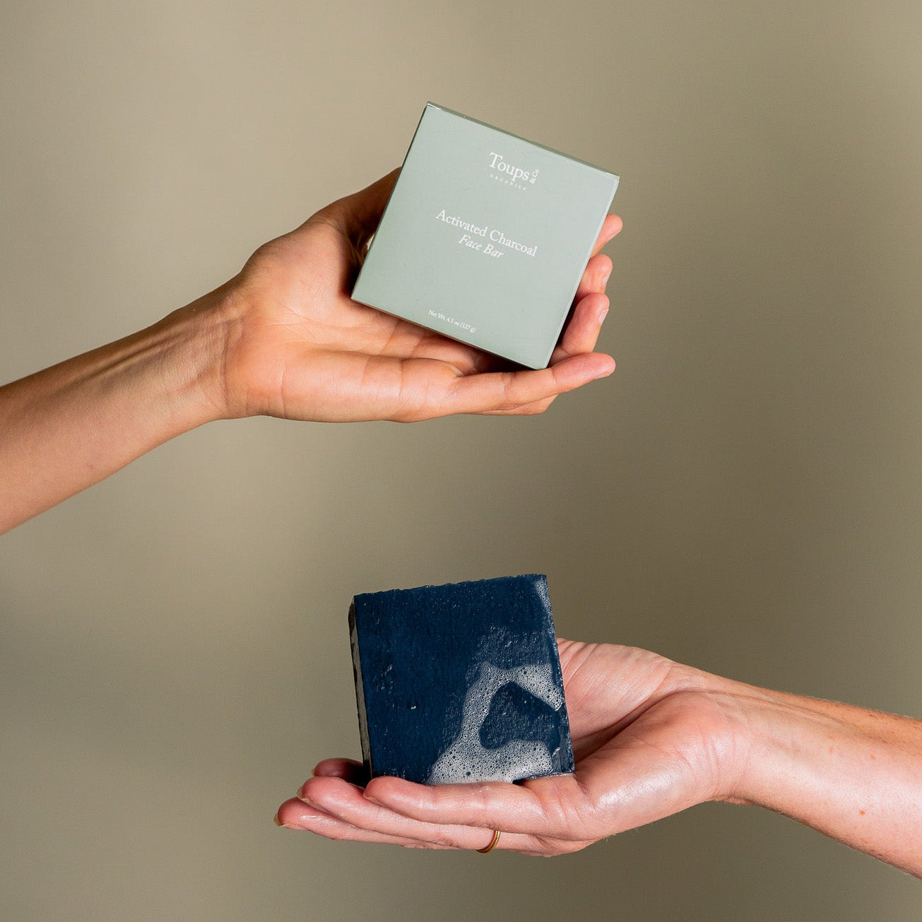 Two hands display a green box labeled “Activated Charcoal Face Bar” from Toups and Co Organics, along with a rectangular dark soap bar marbled with lighter patches—crafted with activated bamboo charcoal and nourishing grass-fed tallow—set against a plain beige background.