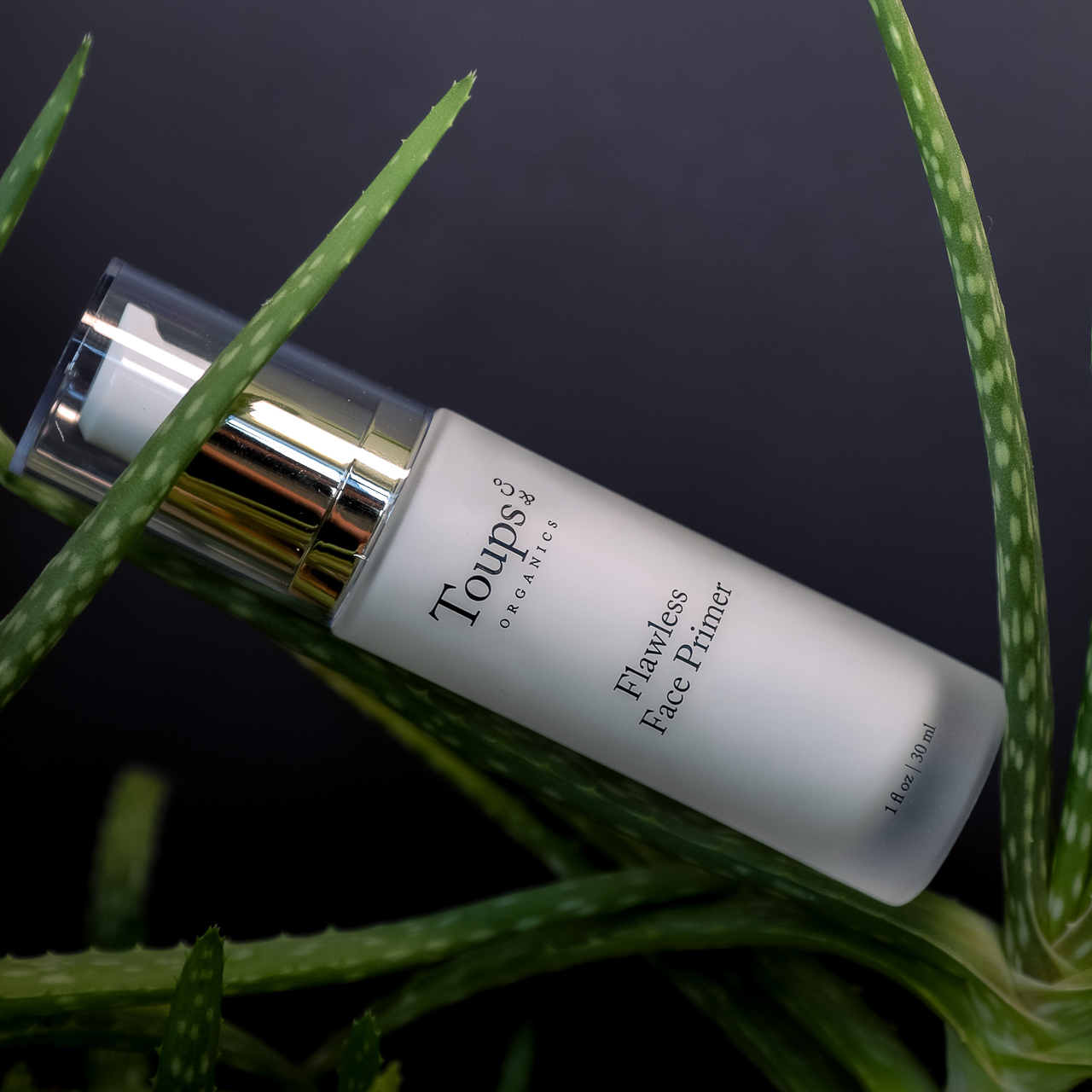 A bottle of Toups and Co Organics Flawless Face Primer, a hydrating primer suitable for all skin types, rests among green aloe vera leaves against a dark background.