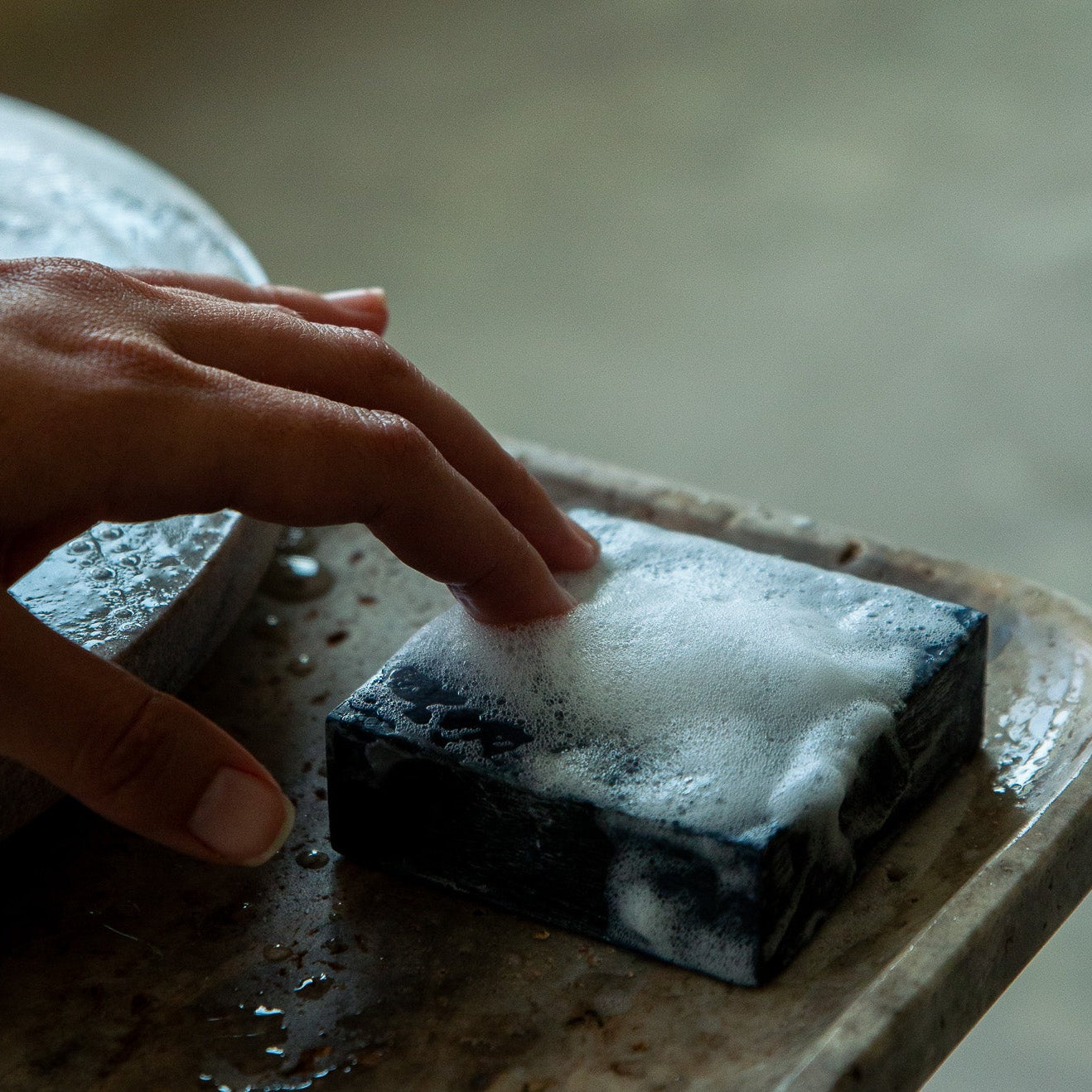 A hand reaches toward the Toups and Co Organics Activated Charcoal Face Bar, crafted with activated bamboo charcoal and covered in white foam, resting on a wet surface.