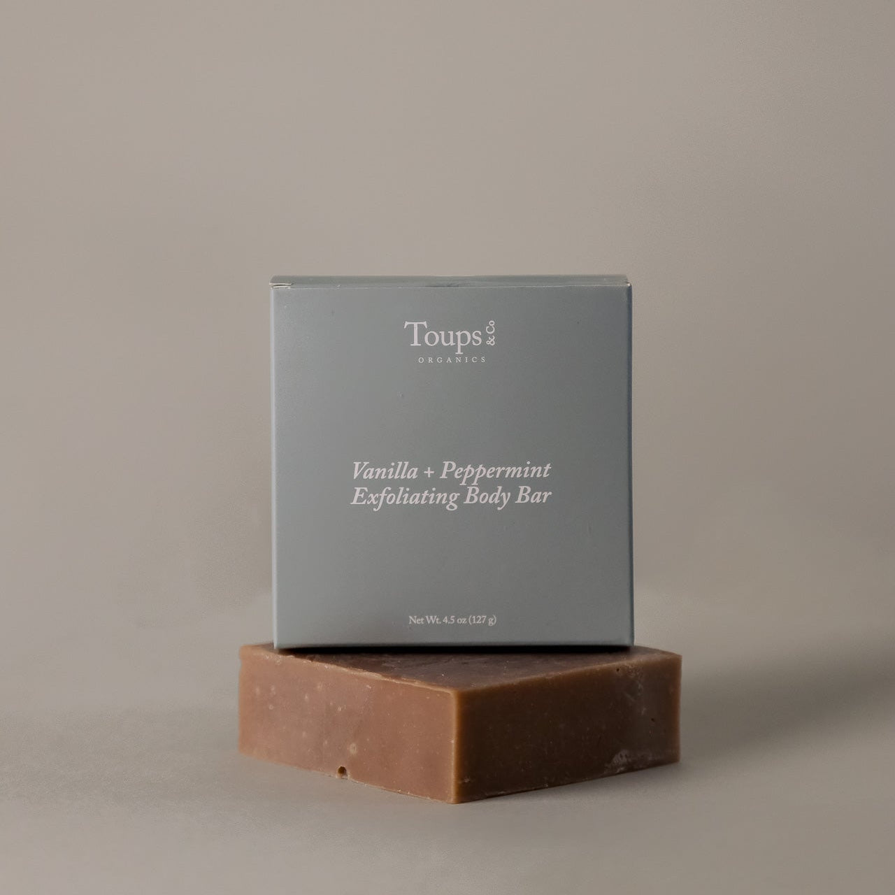 A box labeled "Vanilla Peppermint Exfoliating Body Bar" by Toups and Co Organics rests on top of an unwrapped brown soap bar against a plain background.