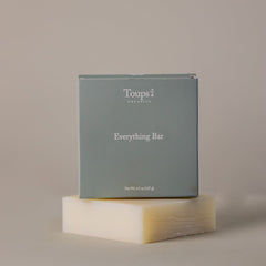 A gray box labeled Toups and Co Organics Everything Bar rests atop an off-white bar made with organic coconut oil, all set against a neutral background.