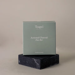 A box labeled Toups and Co Organics Activated Charcoal Face Bar, made with grass-fed tallow and activated bamboo charcoal, sits on a dark stone platform against a plain background.