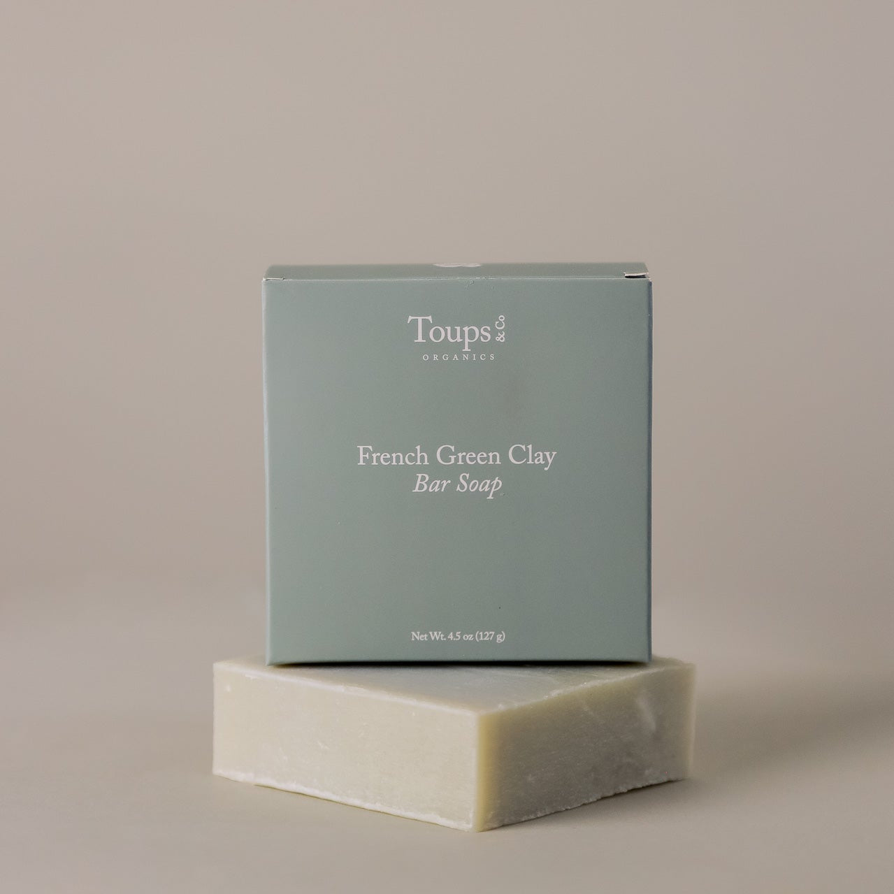 A box labeled "French Green Clay Soap" from Toups and Co Organics rests atop an unwrapped bar of soap against a neutral background, emphasizing its natural skincare benefits and gentle acne control properties.