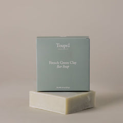 A box labeled "French Green Clay Soap" from Toups and Co Organics rests atop an unwrapped bar of soap against a neutral background, emphasizing its natural skincare benefits and gentle acne control properties.