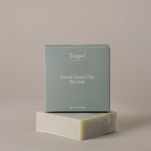 A box labeled "French Green Clay Soap" from Toups and Co Organics rests atop an unwrapped bar of soap against a neutral background, emphasizing its natural skincare benefits and gentle acne control properties.