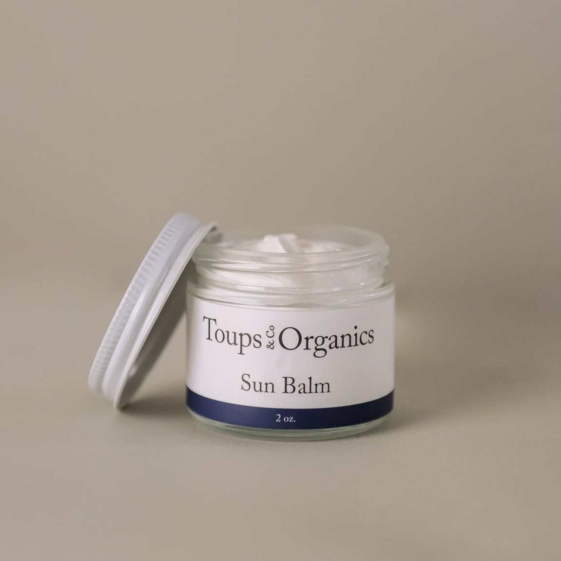 A small glass jar labeled Toups and Co Organics Sun Balm, with a white screw-on lid set aside, holds a white cream formulated with natural mineral sunscreen ingredients such as non-nano zinc oxide.