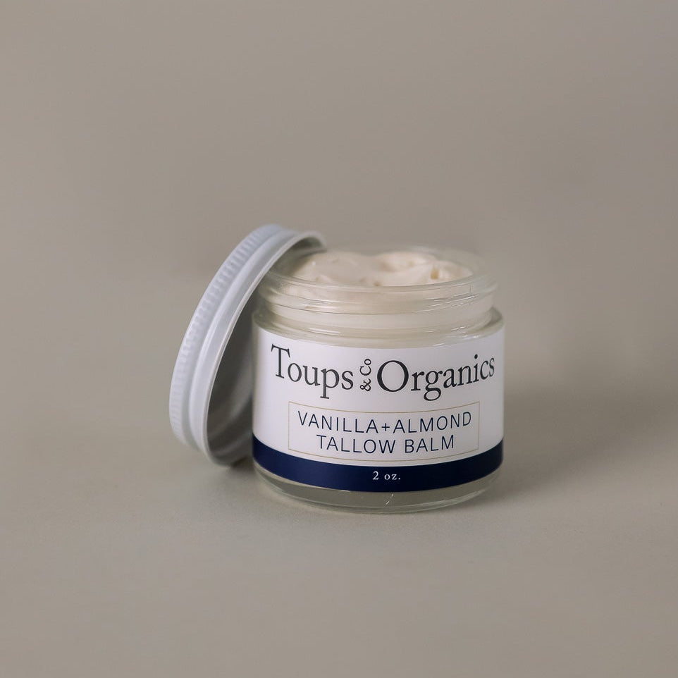 A small glass jar of Toups and Co Organics Tallow Balm - Vanilla + Almond, made with nourishing grass-fed tallow for superior skin hydration, sits with its lid partially open to reveal the creamy balm inside against a plain beige background.