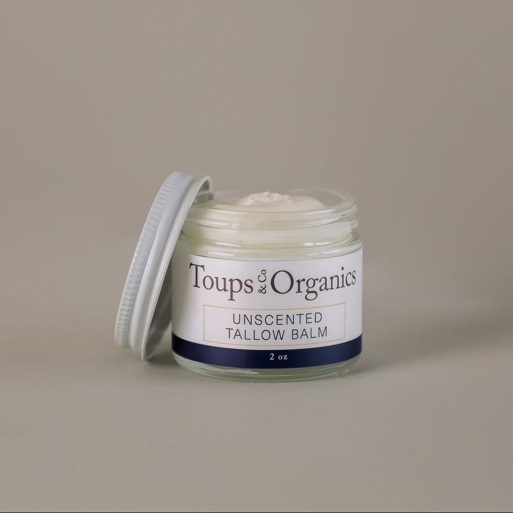 A small glass jar of Toups and Co Organics Tallow Balm - Unscented, featuring nourishing grass-fed tallow and topped with a partially open white screw-on lid, rests on a plain beige background.