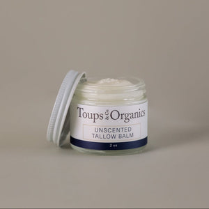 A small glass jar of Toups and Co Organics Tallow Balm - Unscented, featuring nourishing grass-fed tallow and topped with a partially open white screw-on lid, rests on a plain beige background.