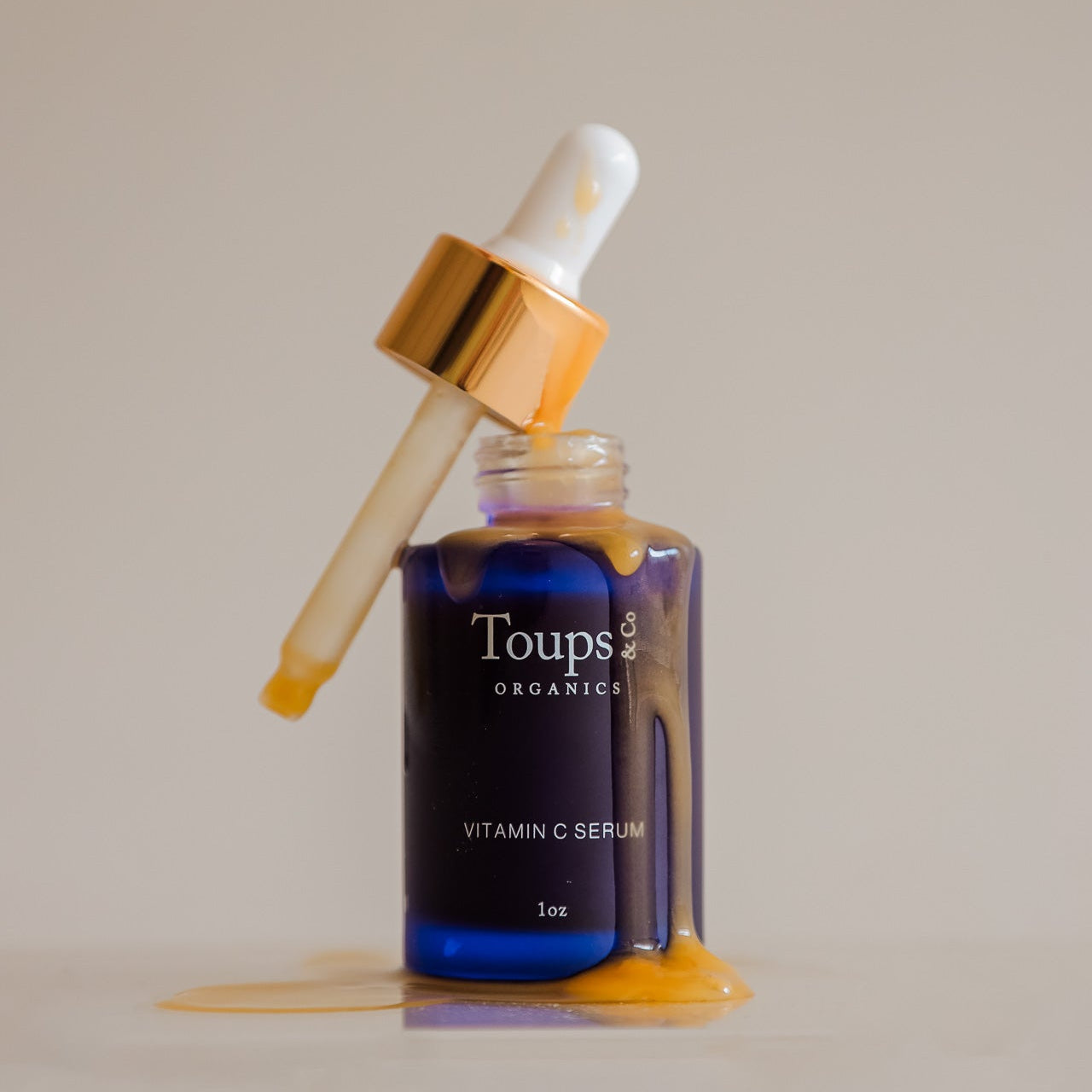 A glass bottle of Toups and Co Organics Vitamin C Serum features a dropper, with serum spilling and dripping down the bottle onto the surface.