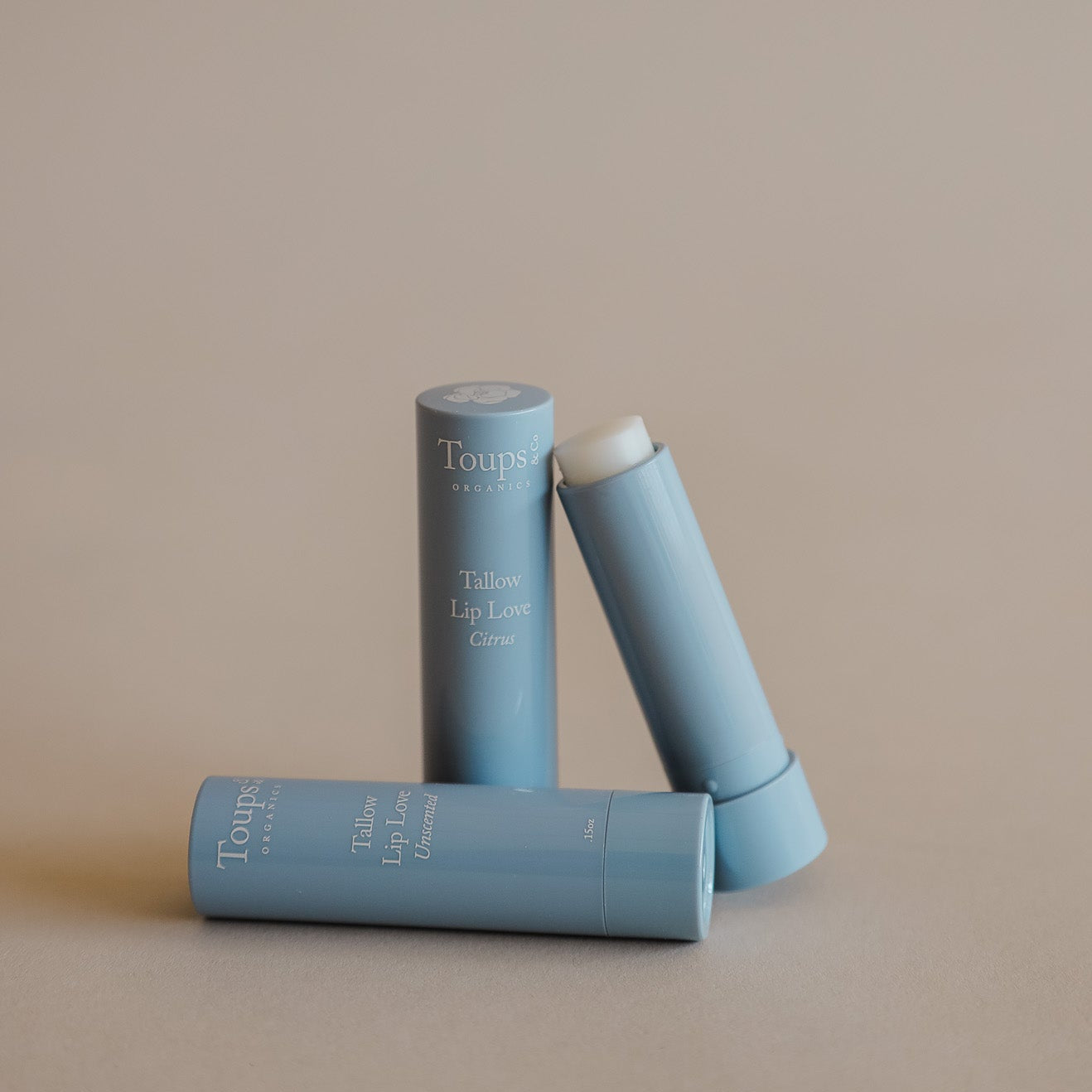 Three blue tubes of Toups and Co Organics Tallow Lip Love lip balm, one open to display the product, arranged on a plain beige background.