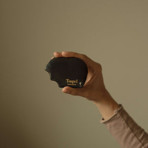 A hand holds a black Nephrite Gua Sha by Toups and Co Organics, ideal for lymphatic drainage, set against a plain beige background.