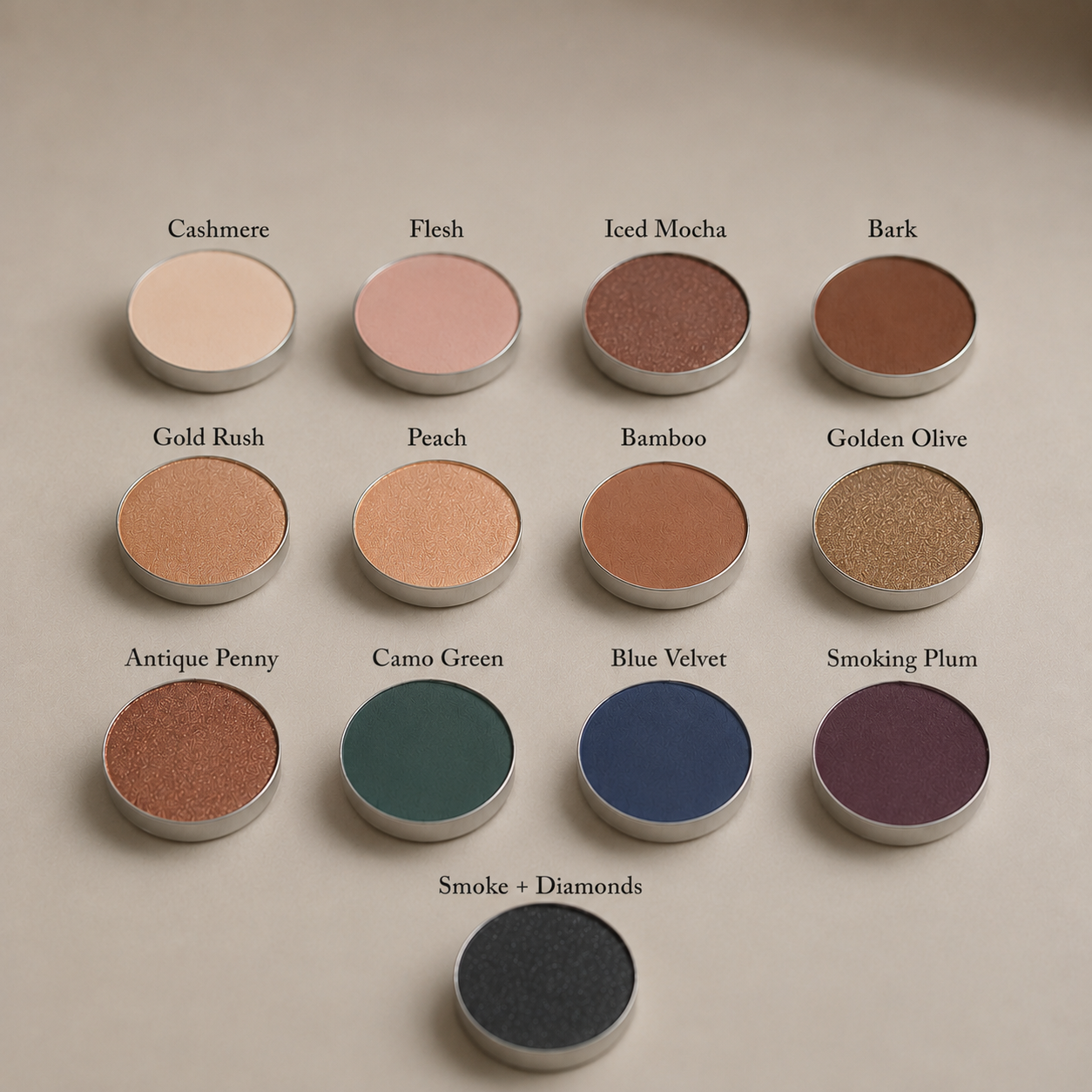 Twelve round, richly pigmented Toups and Co Organics Natural Eyeshadow Singles, each labeled with names in various shades, are arranged in rows on a tan background. This magnetic eyeshadow palette allows you to customize your look with clean, single pans for effortless, vibrant color every time.