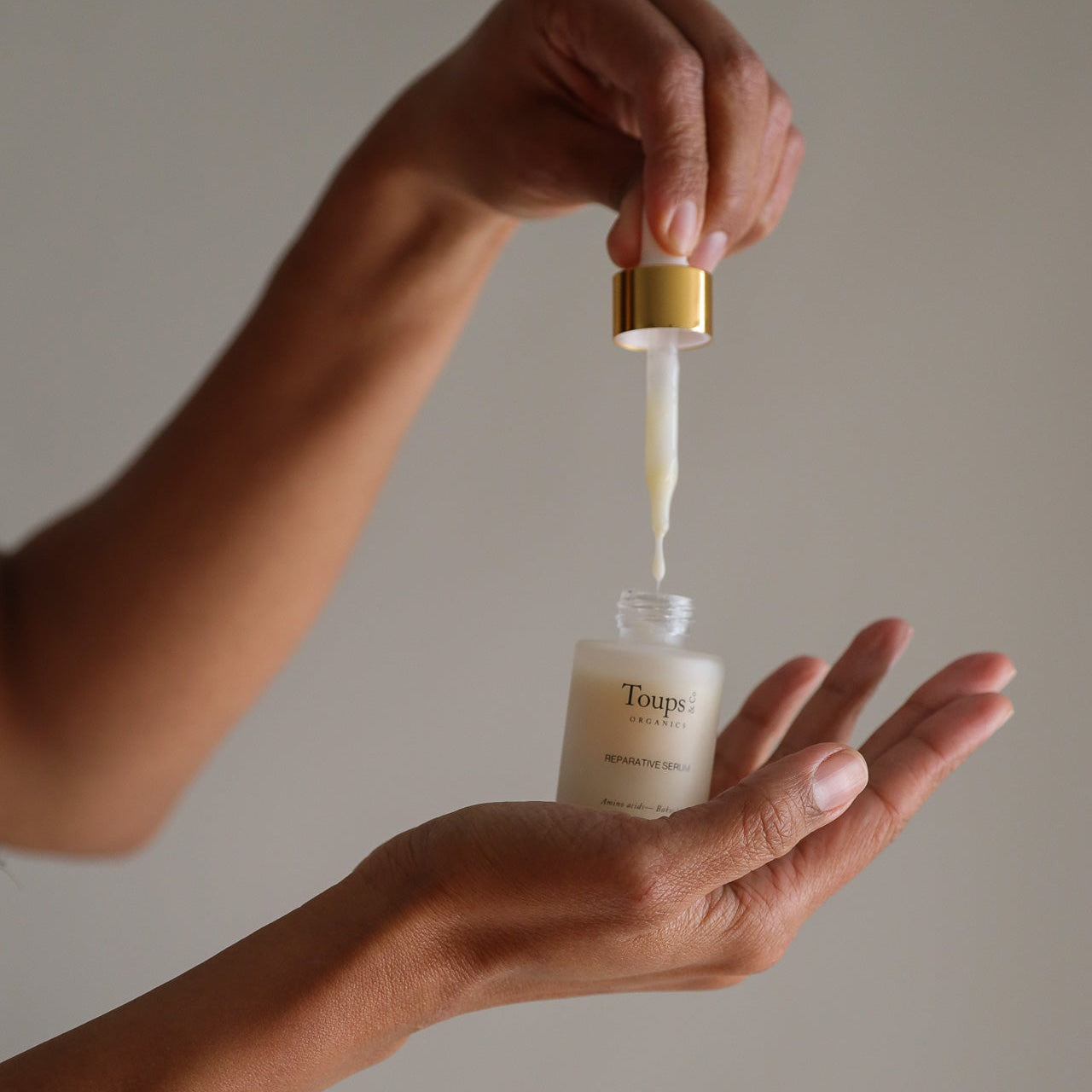 A person holds a dropper above their palm, dispensing serum from a bottle labeled “Toups and Co Organics Reparative Serum”—an anti-aging serum designed for wrinkle prevention—set against a plain background.