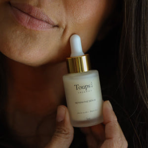 A person holds a bottle of Toups and Co Organics Reparative Serum, using the dropper near their face with a slight smile.