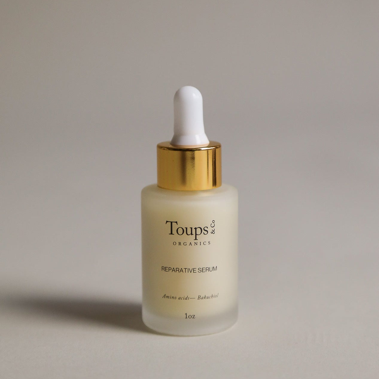 A glass dropper bottle labeled Toups and Co Organics Reparative Serum with a gold cap stands on a neutral background—the ideal anti-aging serum for preventing wrinkles.
