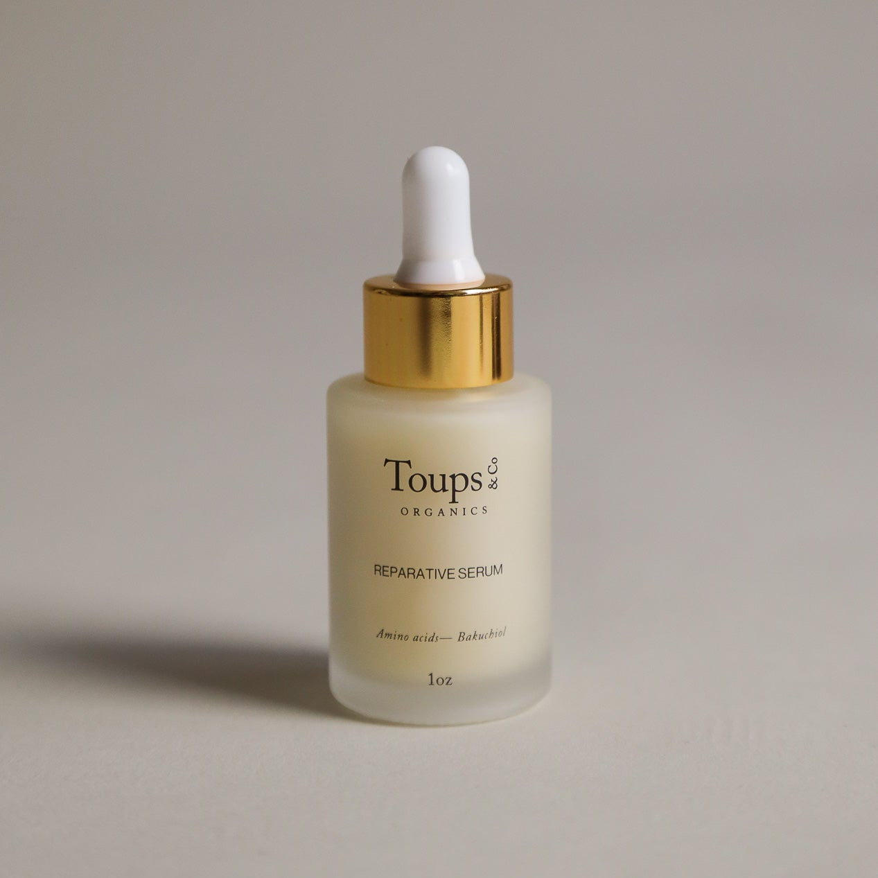 A 1 oz frosted glass dropper bottle labeled Toups and Co Organics Reparative Serum, topped with a gold cap and white bulb, holds a luxurious anti-aging serum formulated with bakuchiol to help prevent wrinkles.