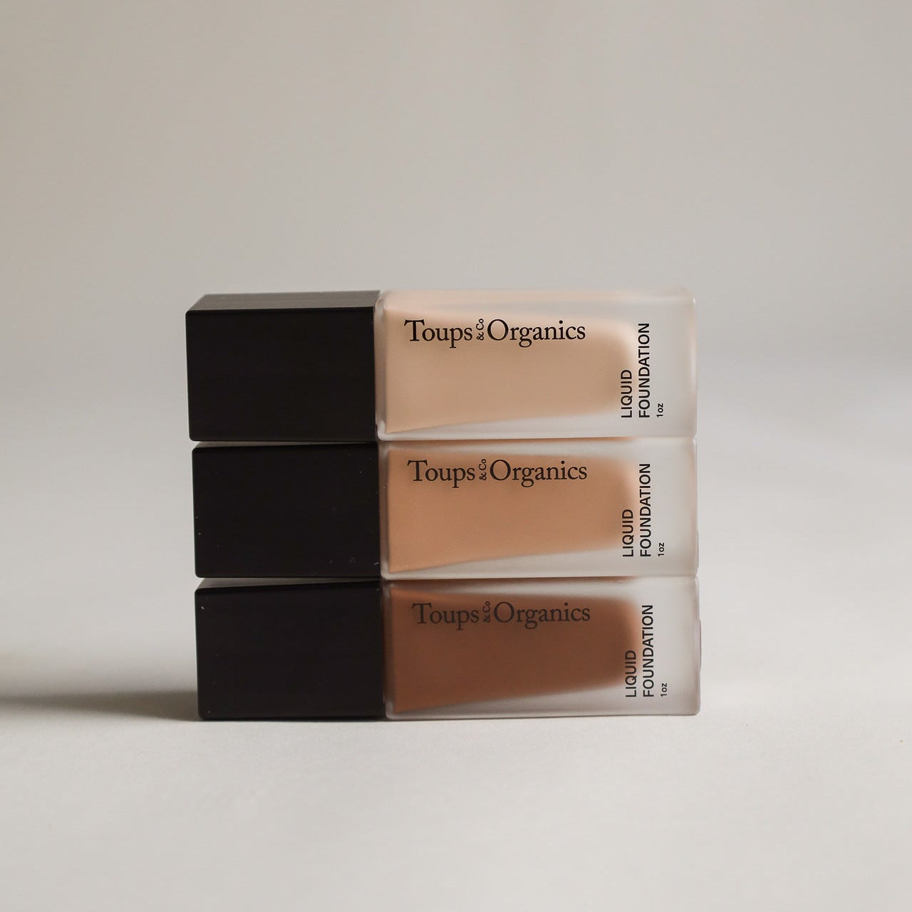 Three rectangular bottles of Toups and Co Organics Liquid Foundation in different shades are stacked horizontally against a simple, neutral background, showcasing their seamless coverage—ideal for sensitive skin.