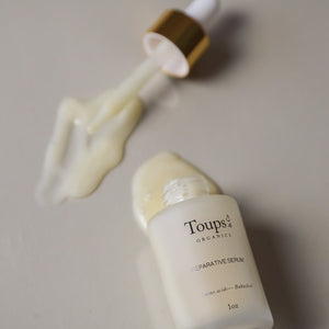 A bottle of Toups and Co Organics Reparative Serum, a nourishing bakuchiol serum formulated for wrinkle prevention, lies on its side with the dropper nearby and a small amount of serum spilled on a smooth surface.