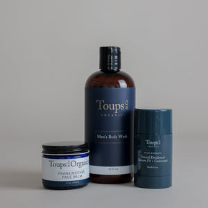 Three Men’s Non Toxic Essentials from Toups and Co Organics—frankincense face balm, men’s body wash, and natural deodorant—are displayed against a plain gray background.
