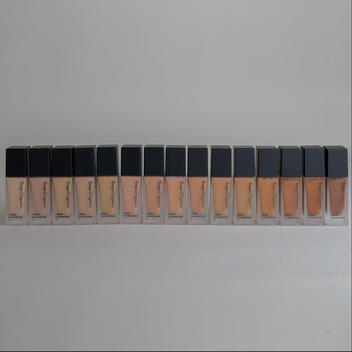 Fifteen bottles of Toups & Co Organics Liquid Foundation arranged in a single row, showing the full range of foundation shades from light to dark, left to right.