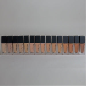 Fifteen bottles of Toups & Co Organics Liquid Foundation arranged in a single row, showing the full range of foundation shades from light to dark, left to right.