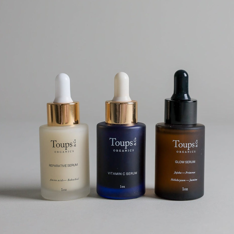 Three Toups and Co Organics Clean Serum Set bottles with droppers are displayed side by side on a neutral background, featuring labels for Reparative Serum, Vitamin C Serum, and Glow Serum.