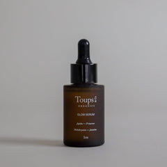 A 1oz brown glass dropper bottle labeled “Glow Serum” by Toups and Co Organics, highlighting vitamin-rich oils and ingredients, displayed against a plain beige background.