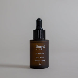 A 1oz brown glass dropper bottle labeled “Glow Serum” by Toups and Co Organics, highlighting vitamin-rich oils and ingredients, displayed against a plain beige background.