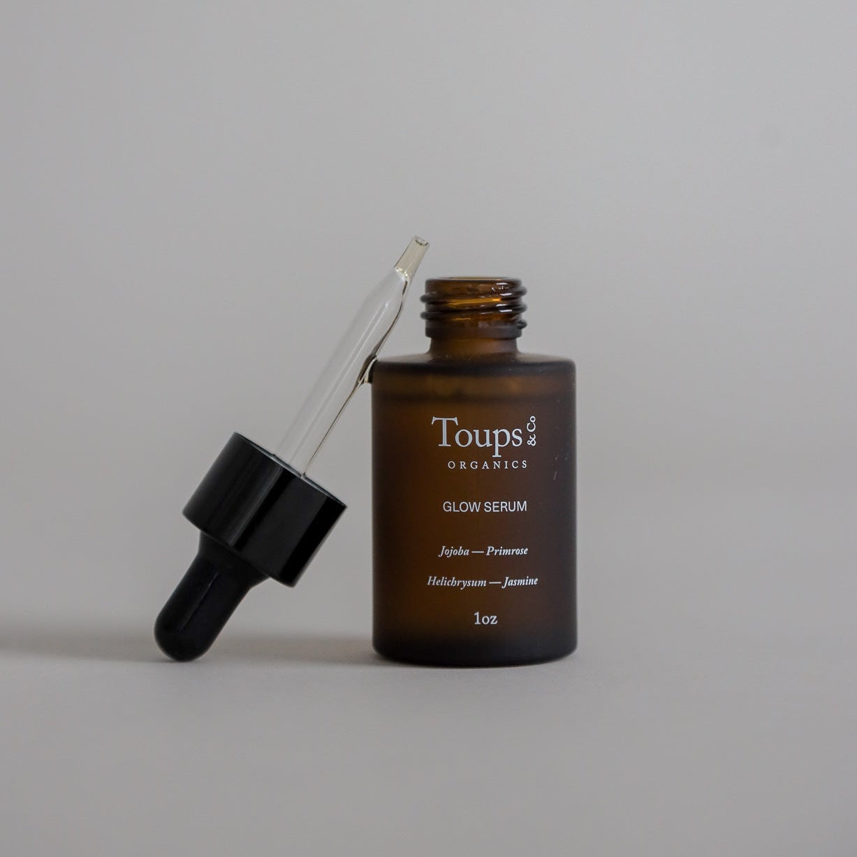 A 1 oz brown glass dropper bottle labeled "Glow Serum" by Toups & Co Organics with the dropper resting on top of the open bottle, displayed on a plain beige background to highlight its vitamin-rich oil blend.