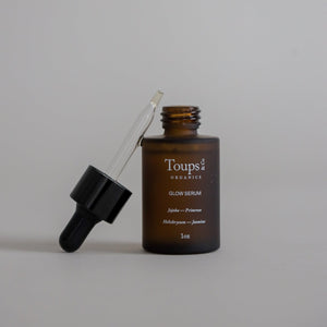 A 1 oz brown glass dropper bottle labeled "Glow Serum" by Toups & Co Organics with the dropper resting on top of the open bottle, displayed on a plain beige background to highlight its vitamin-rich oil blend.