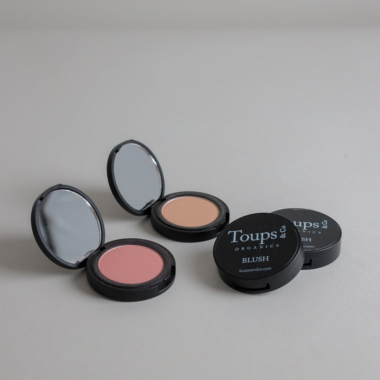 Two open Toups and Co Organics Complete Makeup Set blush compacts, one pink and one beige, displayed on a plain gray surface beside their matching closed lids.