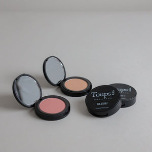Two open Toups and Co Organics Complete Makeup Set blush compacts, one pink and one beige, displayed on a plain gray surface beside their matching closed lids.