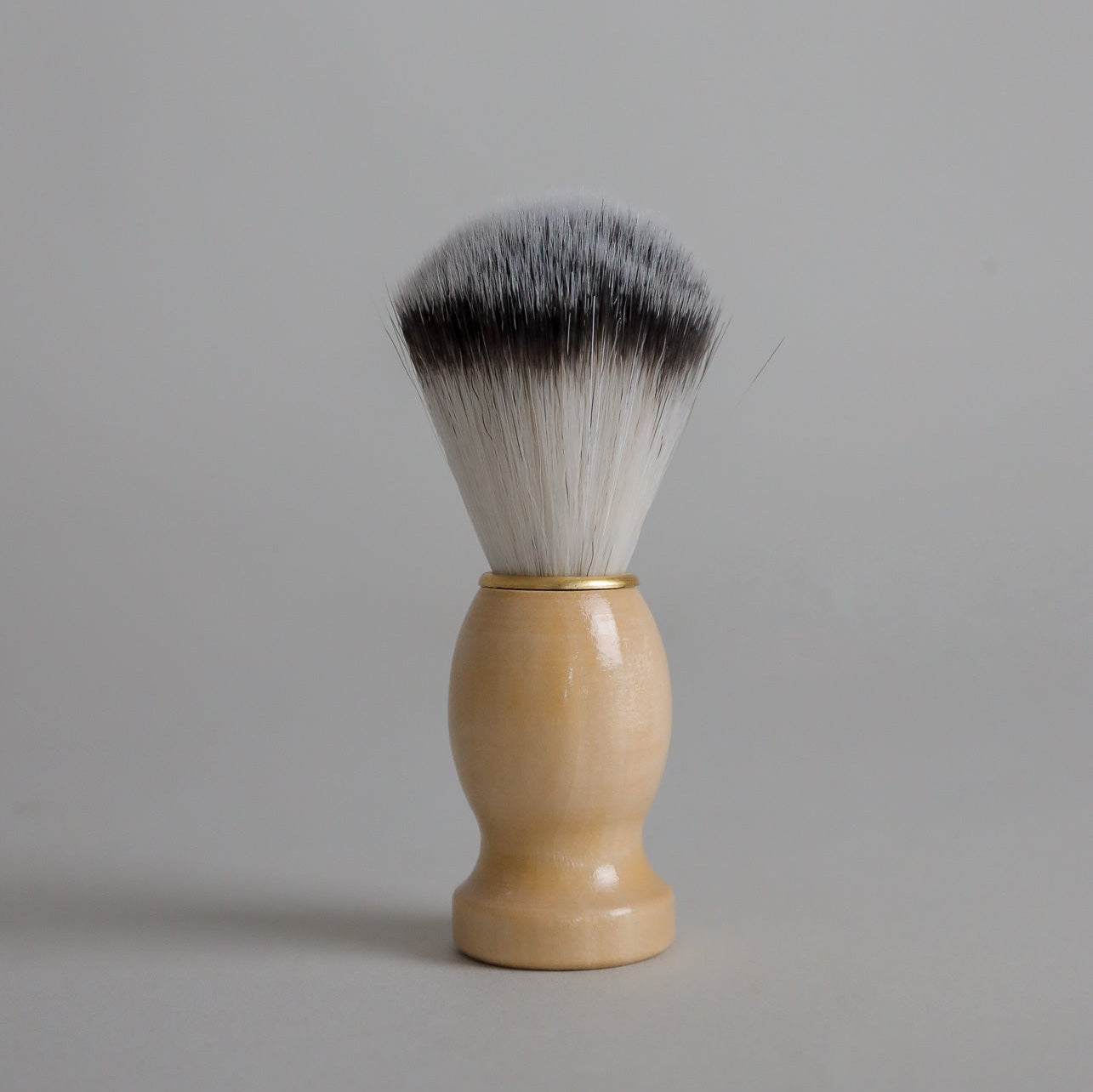 A Toups and Co Organics Men’s Shave Brush, featuring a light wooden handle and soft, two-toned bristles, stands upright against a plain gray background.