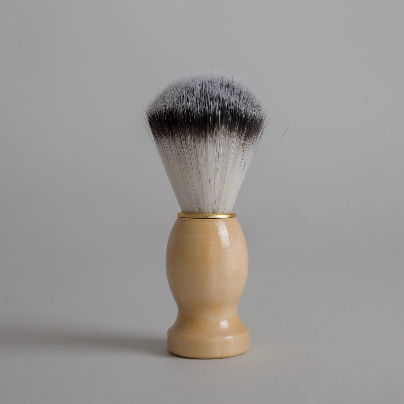 A Toups and Co Organics Men’s Shave Brush, featuring a light wooden handle and soft, two-toned bristles, stands upright against a plain gray background.