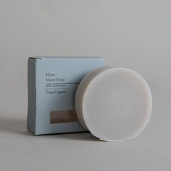 A round bar of white men’s shave soap, crafted as a natural shaving soap, is displayed in front of its light blue box packaging labeled Men’s Shave Soap by Toups and Co Organics.