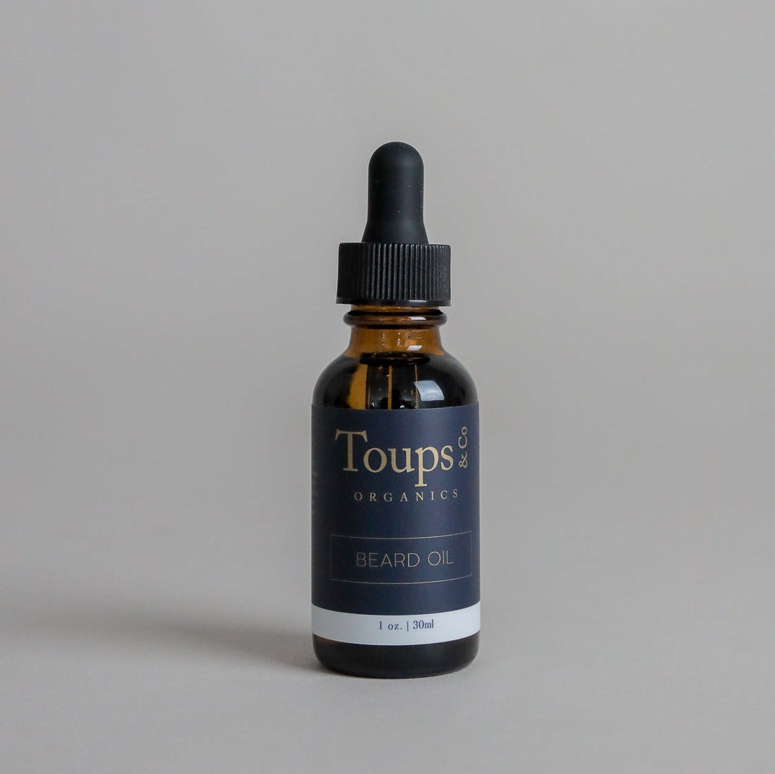 A brown glass bottle of Toups and Co Organics Beard Oil, formulated to promote beard growth and moisturize facial hair, is set against a simple gray background. The black dropper cap provides an elegant accent to this must-have grooming product.